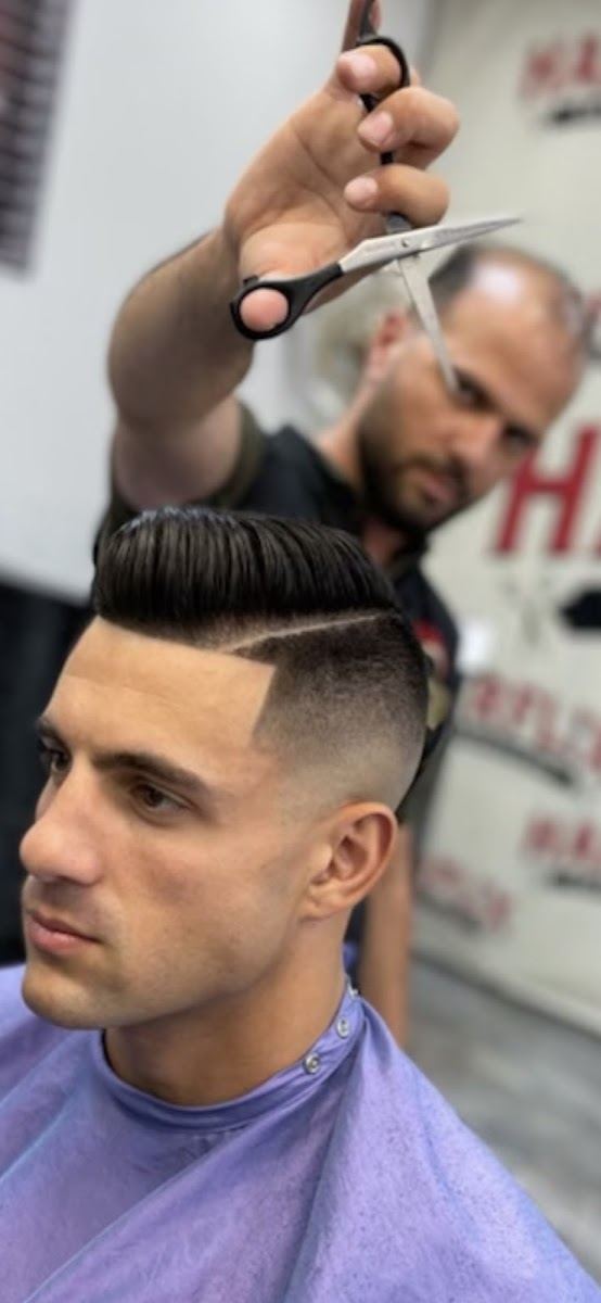 Barber holding scissors over a client's slicked back pompadour with a sharp skin fade and hard part