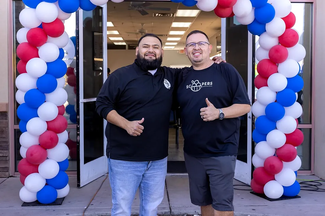 Two men smiling at the shop door under a red, white and blue balloon arch