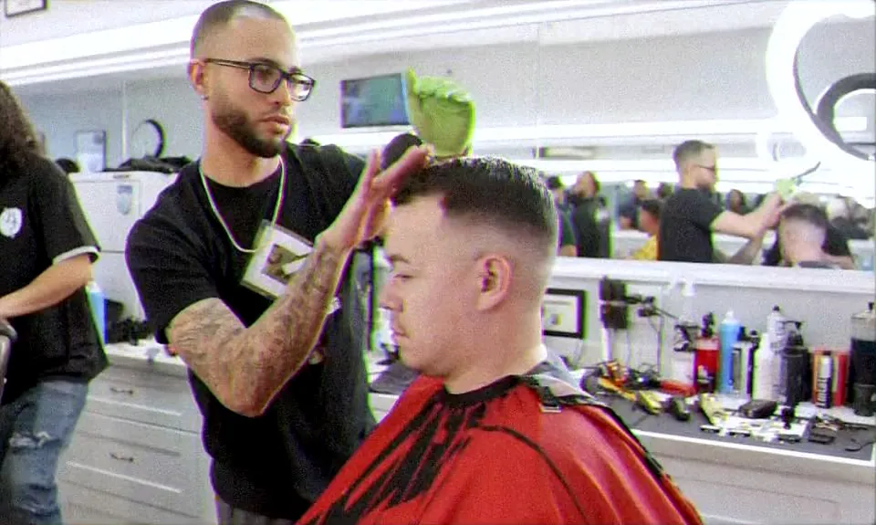 A barber in a black tee shaping a fresh taper on a client under a red cape, mirrors and stations behind