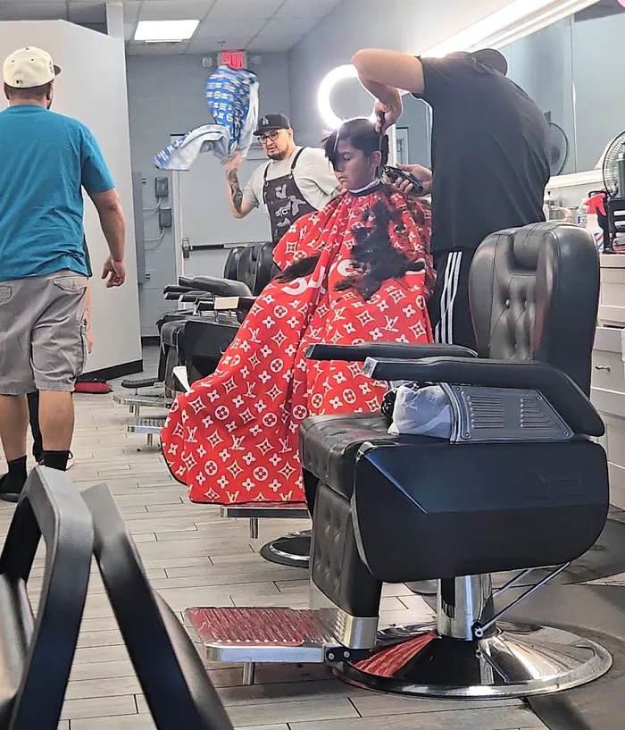 A young boy in a black barber chair under a red cape while a barber works on his hair