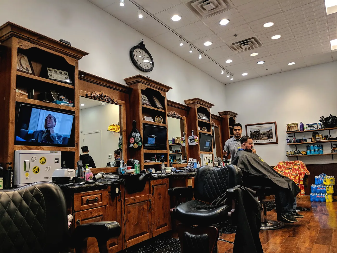 The room: a row of walnut barber stations with mirrors and shelves, black tufted chairs, a wood floor and a large wall clock