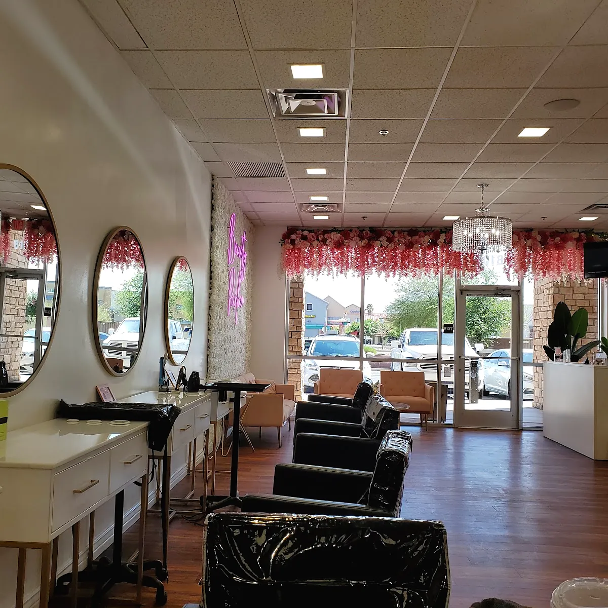 Inside the salon: round mirrors along a white wall, a pink neon script sign on a flower wall, black styling chairs, a chandelier and a pink floral garland over the front windows