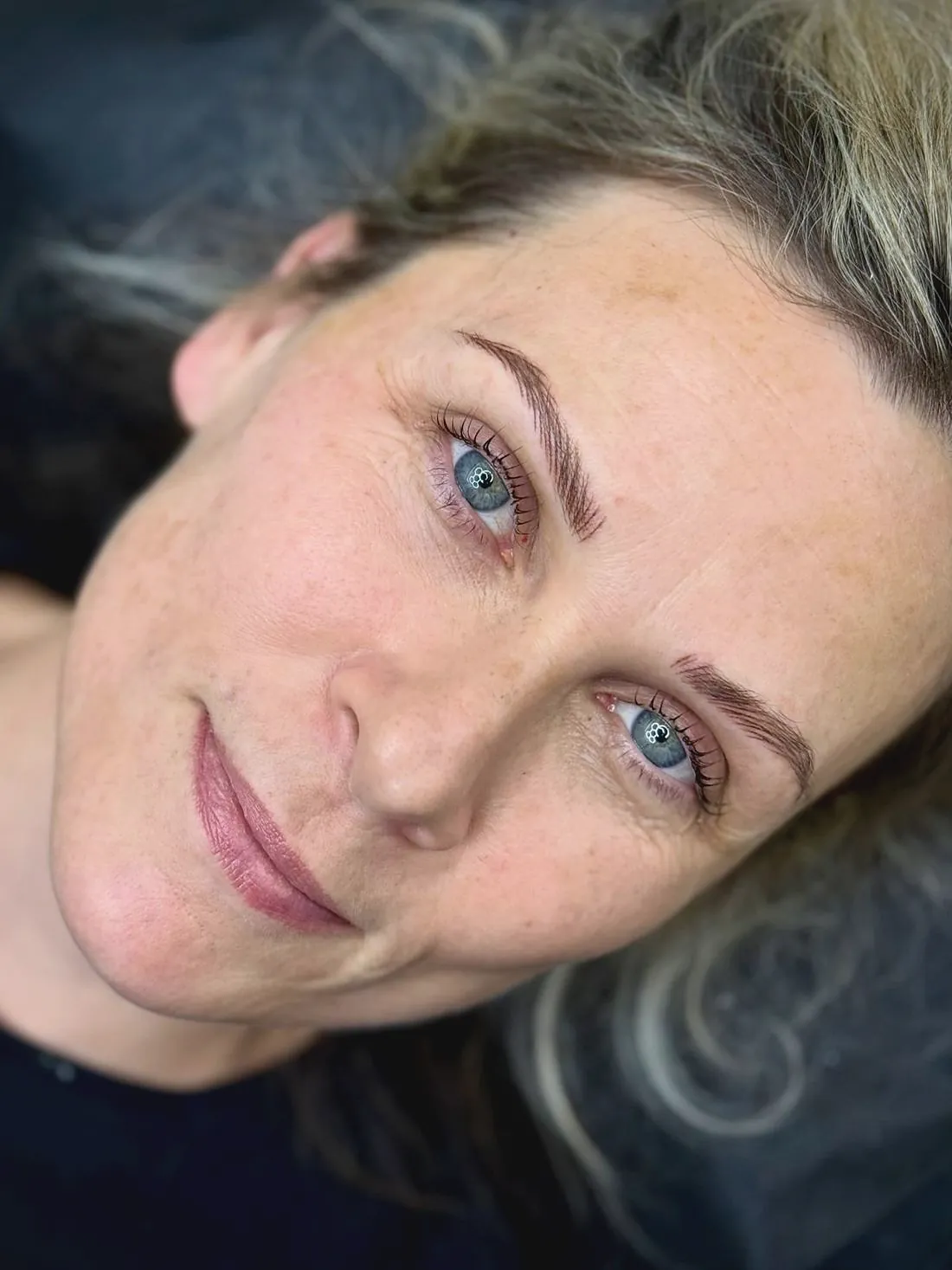 A woman with silver blond hair lying back, soft defined hair stroke brows above blue eyes