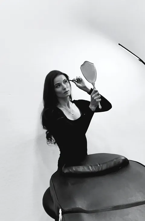 Black and white photo: a woman in black holds up a hand mirror beside a padded treatment bed against a white wall