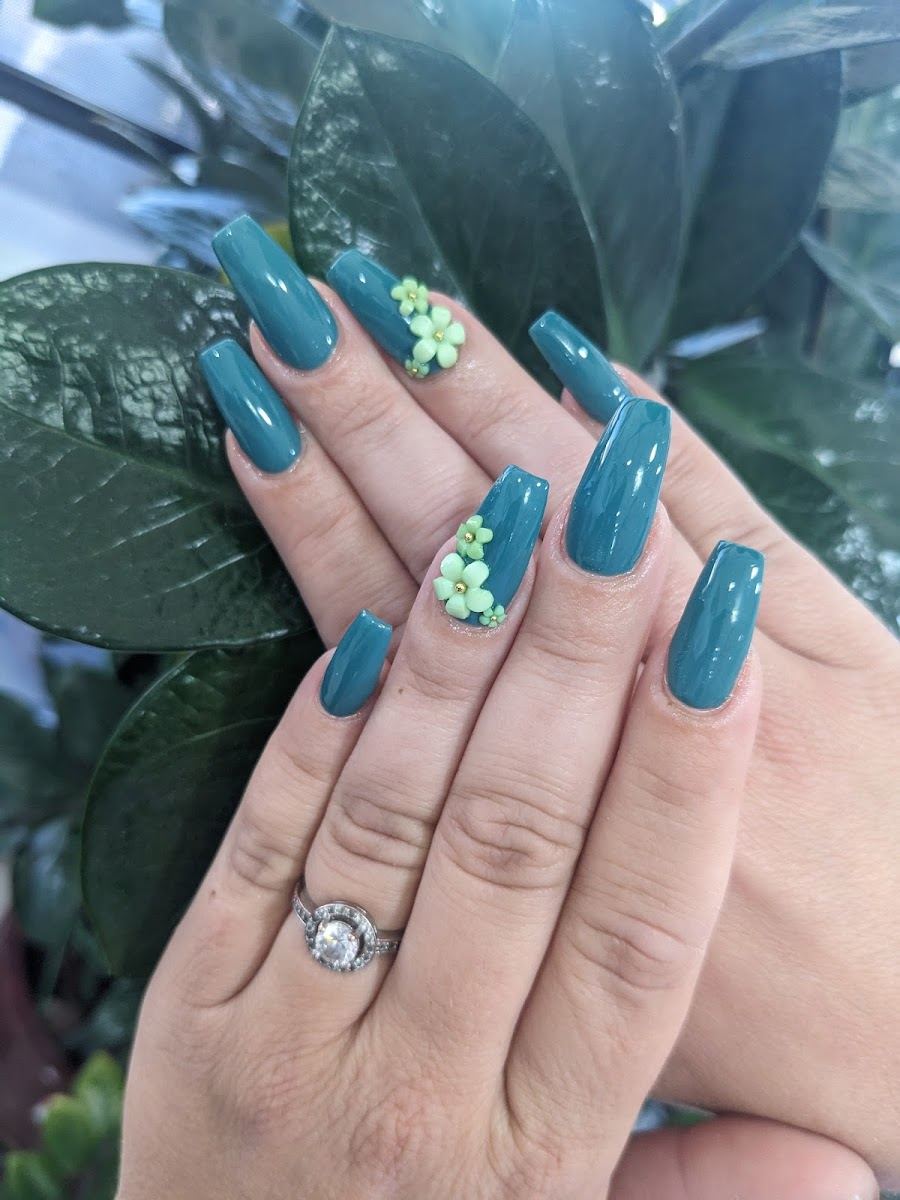 Teal coffin nails with 3D green flower accents, held against leafy plants