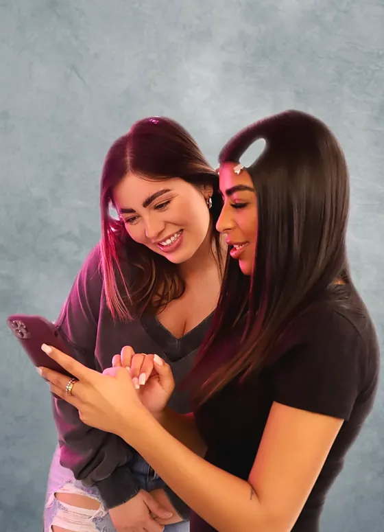 Two women laughing over a phone against a grey backdrop