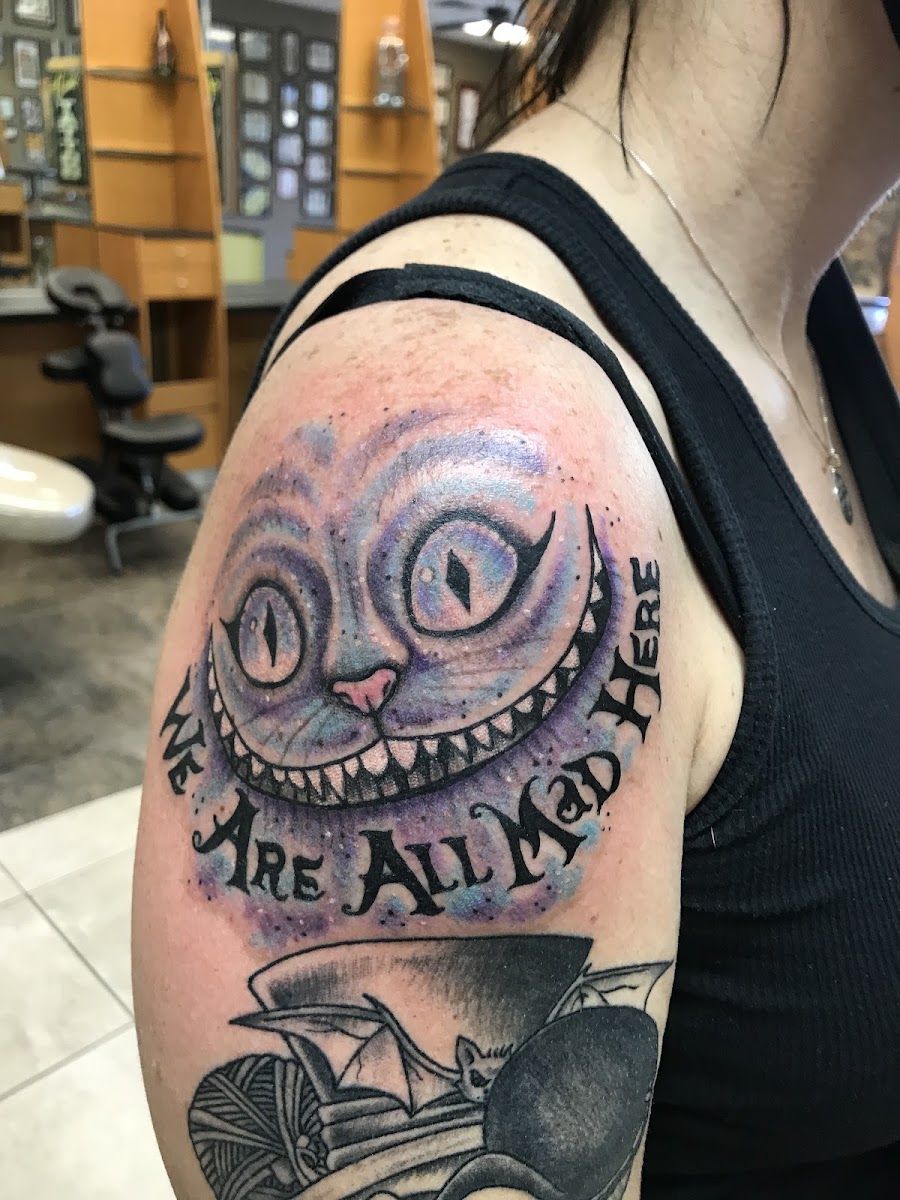 Grinning Cheshire cat tattoo on a shoulder with script lettering