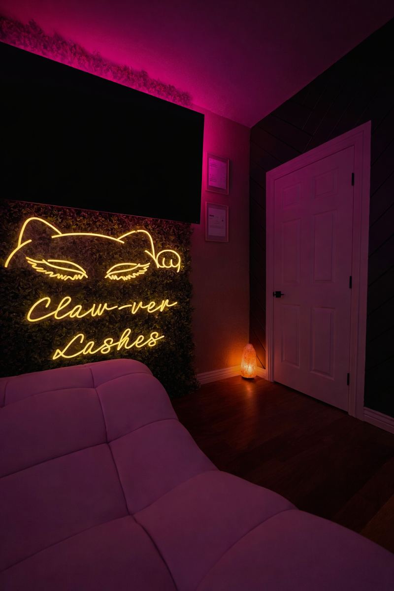 The glowing yellow Claw-ver Lashes neon cat sign above the lash bed in Danae's studio