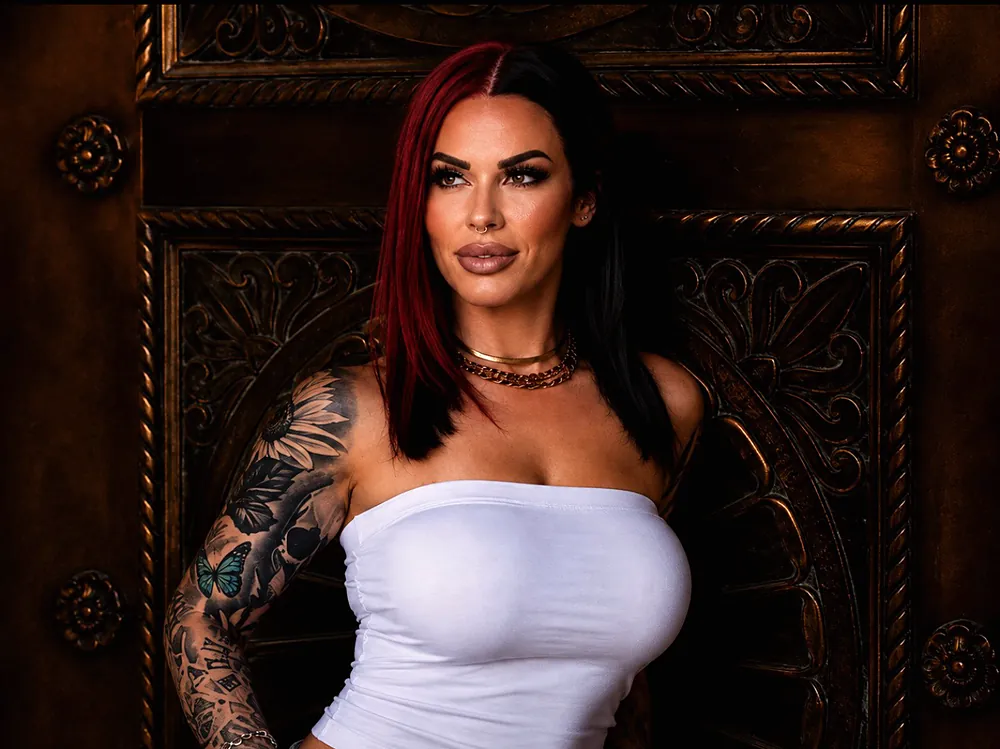 Becca, red and black split bob and tattoo sleeves, in front of a dark carved wooden door