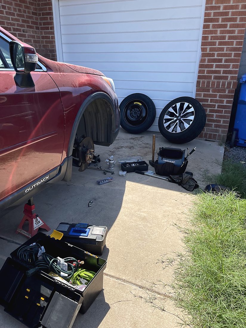 A red SUV with a wheel off on a jack stand beside a garage, two wheels and a toolbox nearby