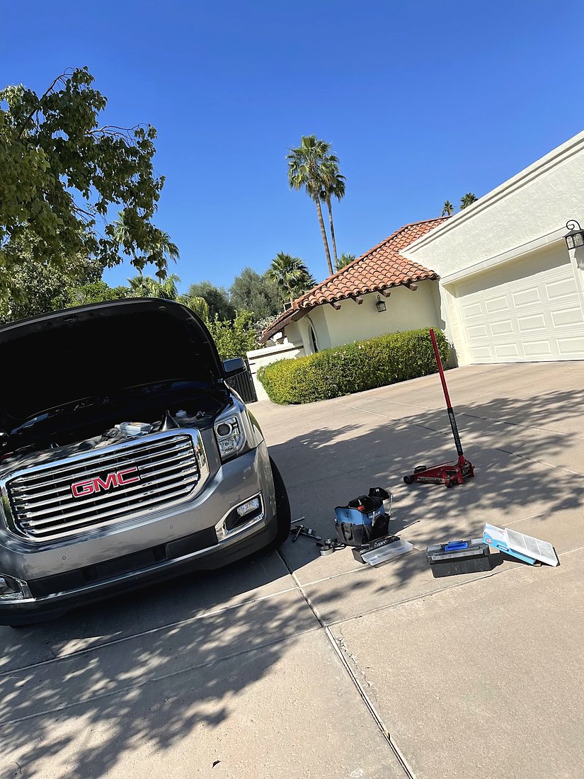 A silver GMC SUV with its hood up in a driveway, parts and boxes on the ground