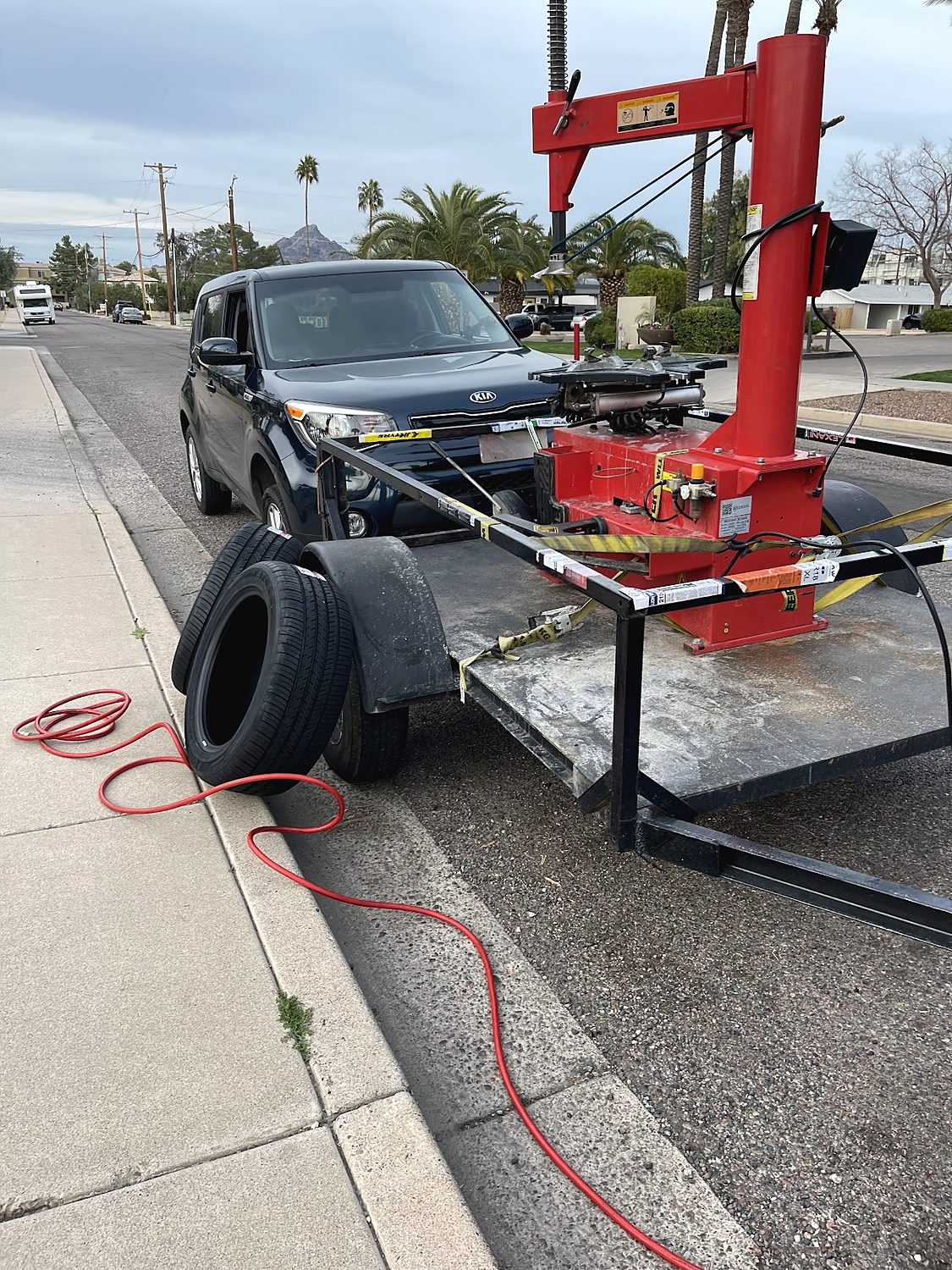 A red tire changing machine and tires on a trailer parked at a curb, a pickup truck behind it