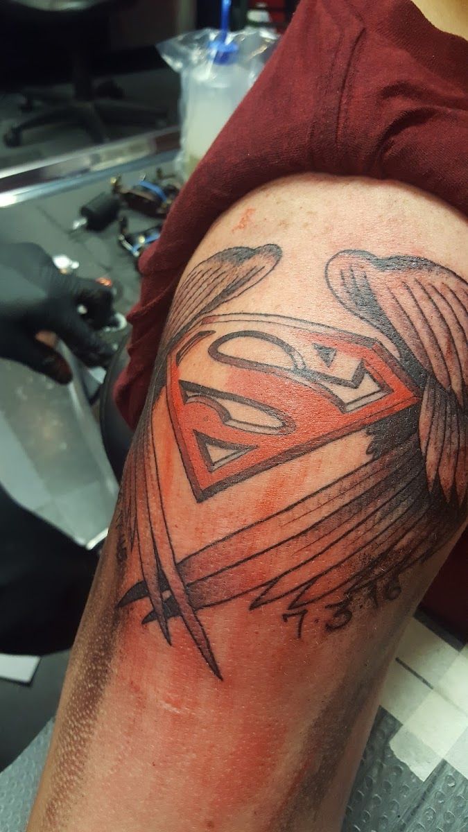Red Superman shield tattoo with a black wing on a shoulder
