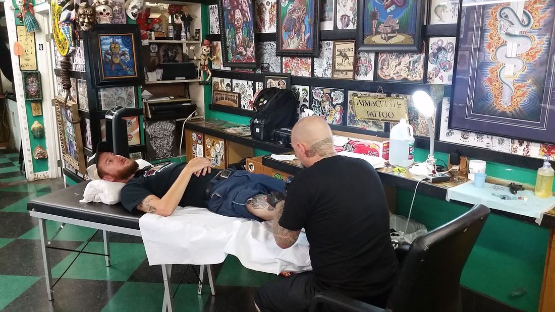 An artist tattooing a client lying on the table under walls of framed flash