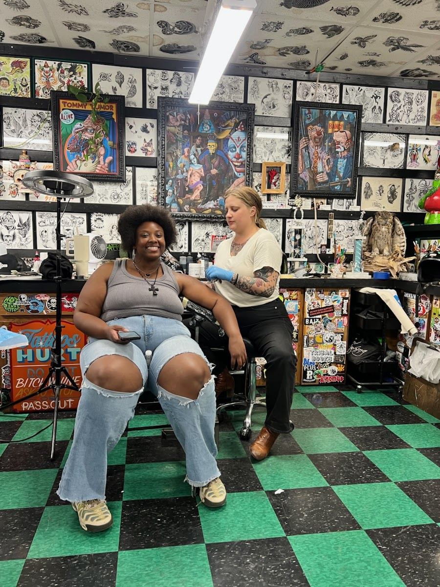 A smiling client seated in the studio next to her artist, teal walls and checkered floor behind