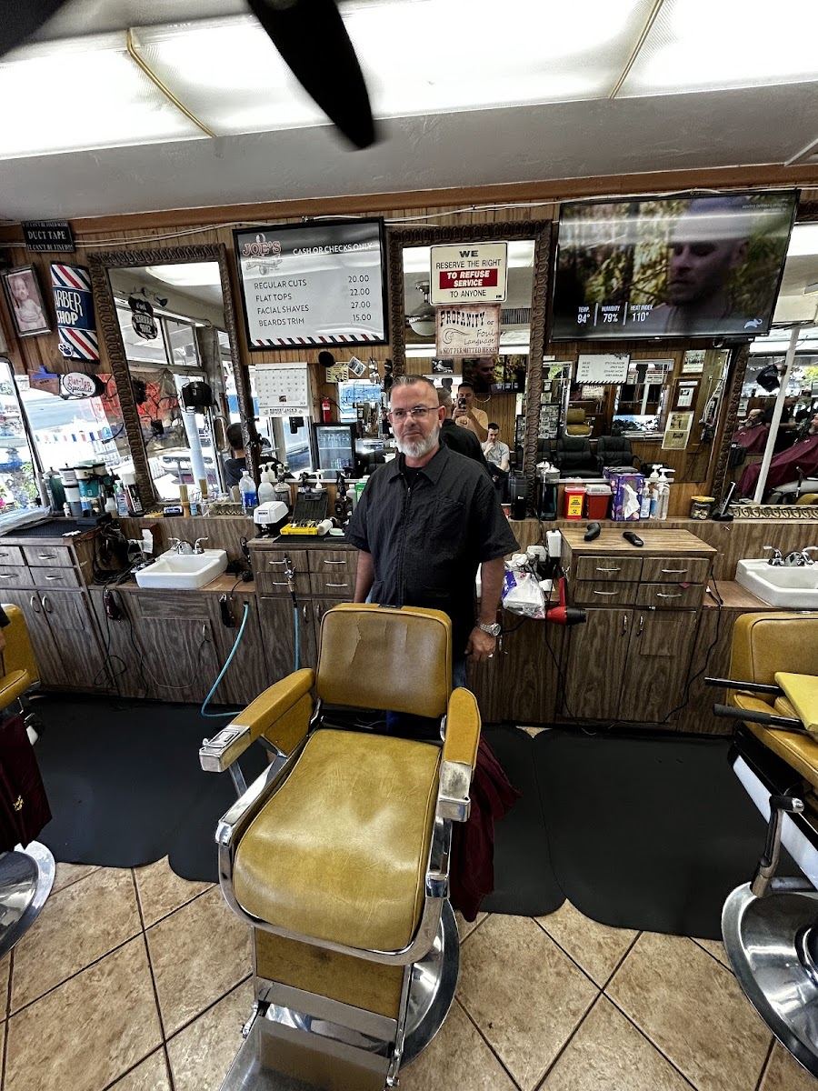 A barber standing behind a gold vinyl barber chair inside the wood-paneled shop