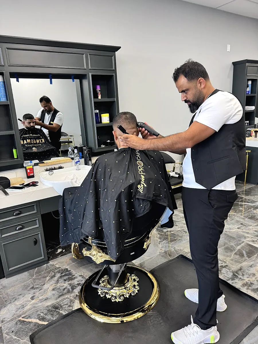 A barber in a black vest cutting a client's hair at a gold and black chair, the client in a black Kingsman cape
