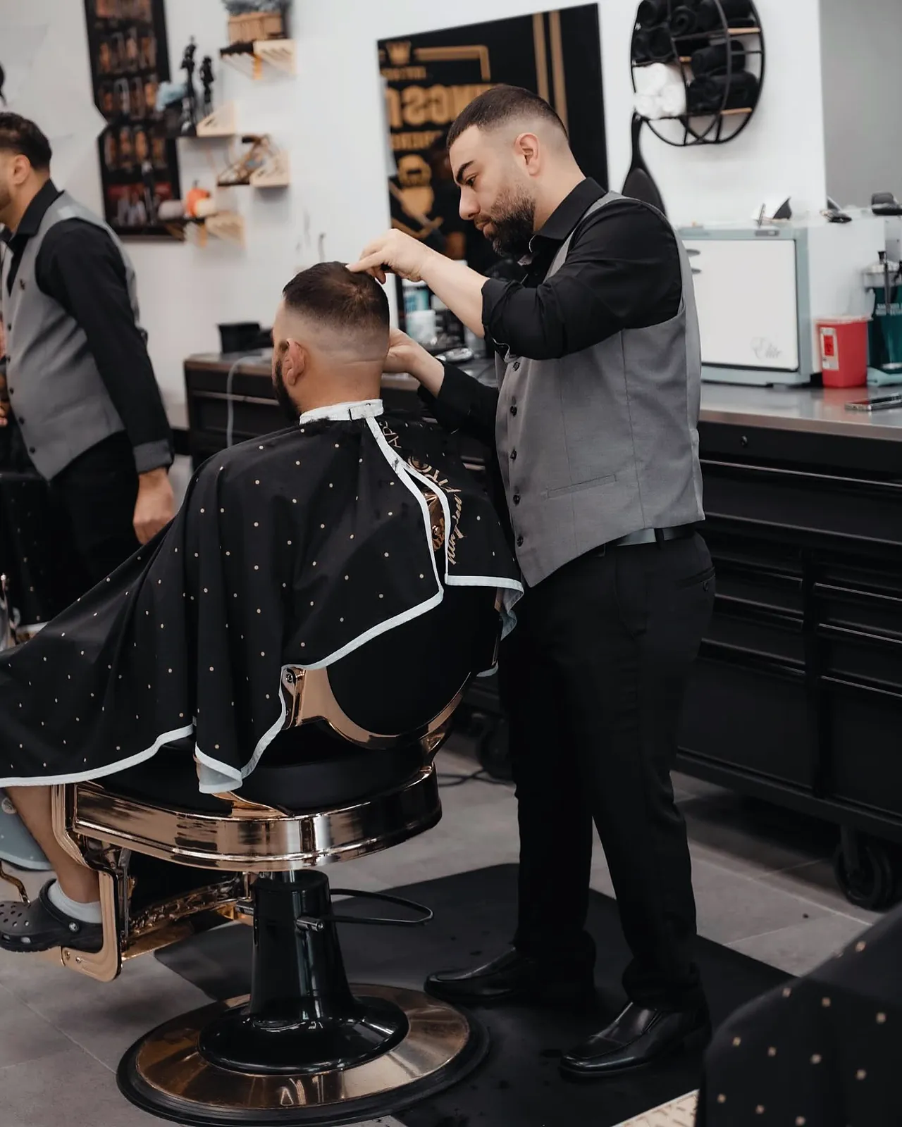 Two barbers in grey vests cutting a client's hair under the Kingsman sign