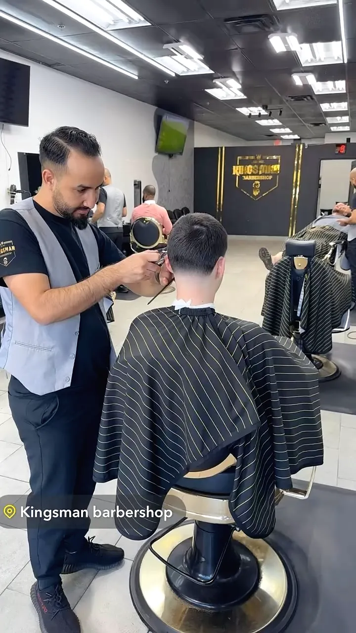 A barber in a grey vest cutting a client's hair at a gold chair
