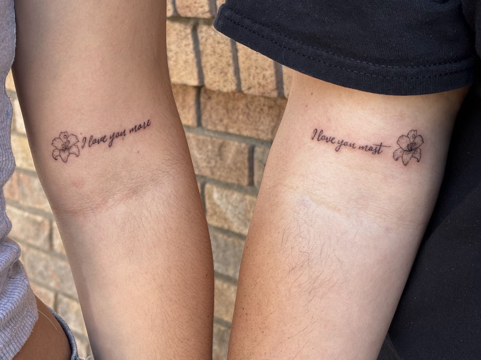 Two forearms with matching fine line script tattoos reading I love you more