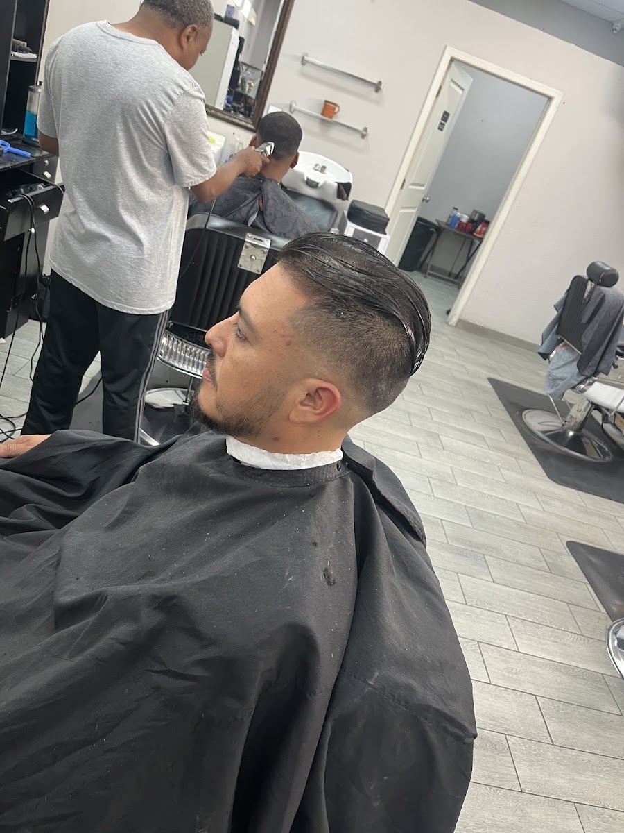 A barber at work in the shop while a client in a black cape shows a fresh taper with a long top