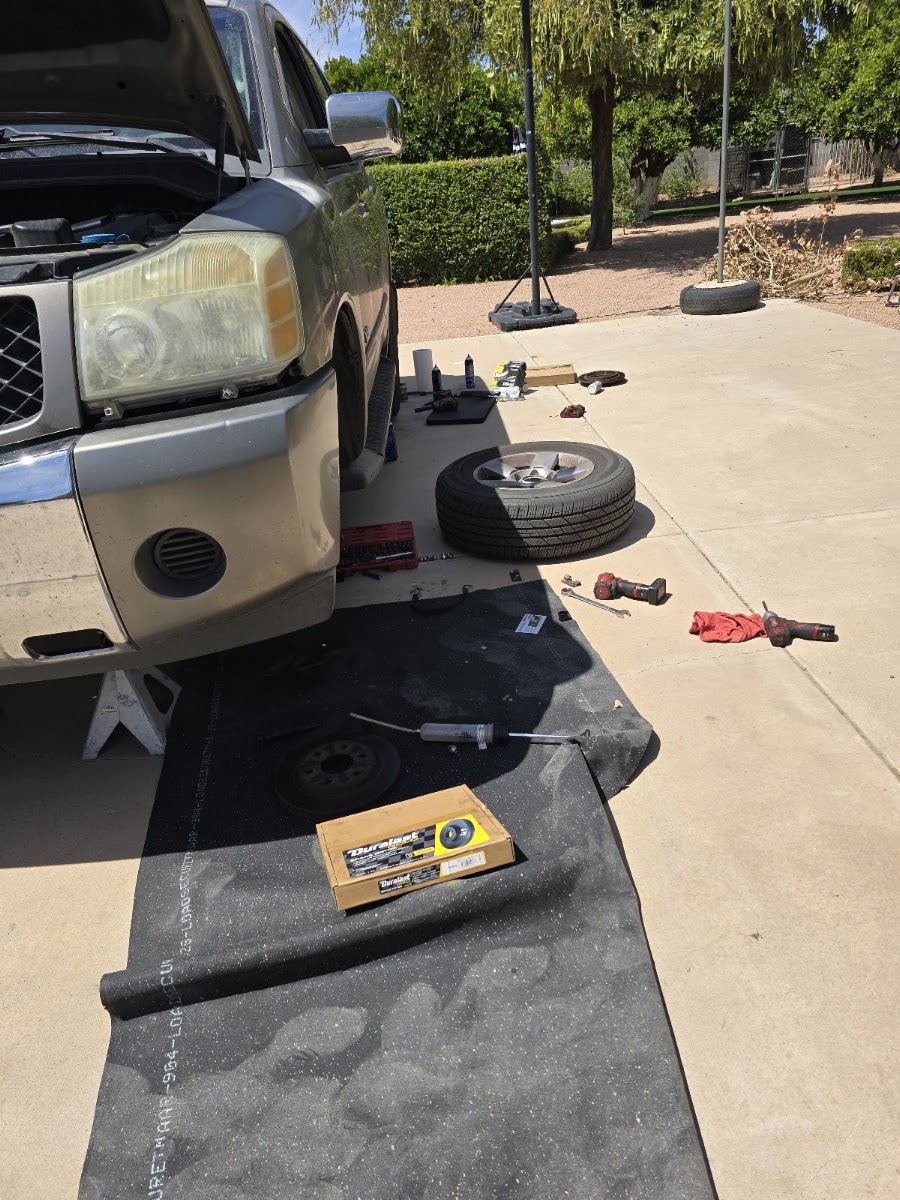 A pickup on jack stands in a sunny driveway with a wheel off, new brake parts and tools laid out on a mat