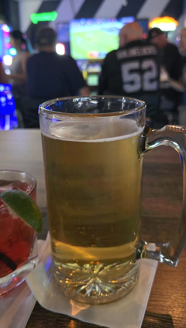 A full glass mug of light beer on a napkin, the bar and a TV behind it