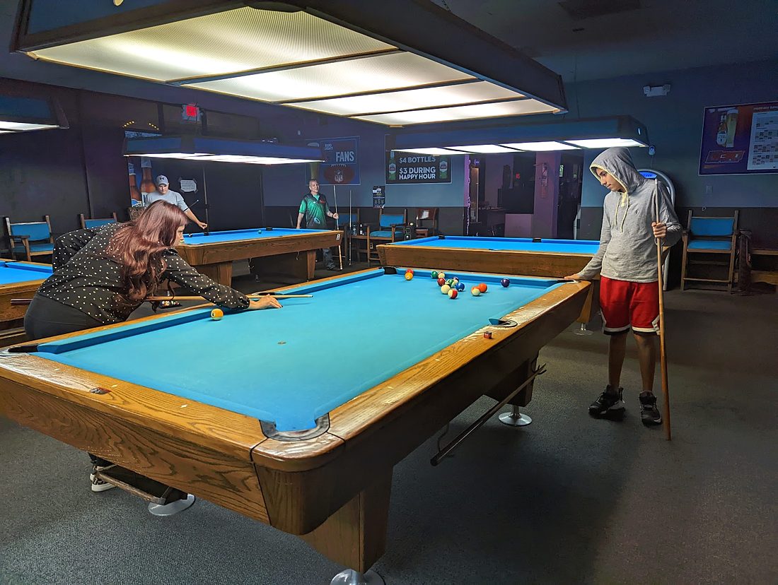 A row of blue-felt pool tables under long lights, two people mid-game at the nearest table