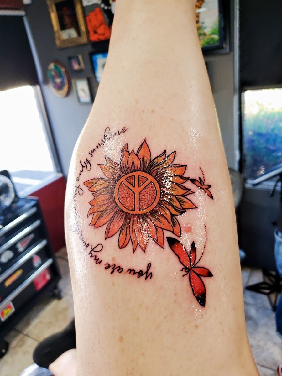 Orange sunflower tattoo with a peace sign center, red dragonfly and script on a forearm