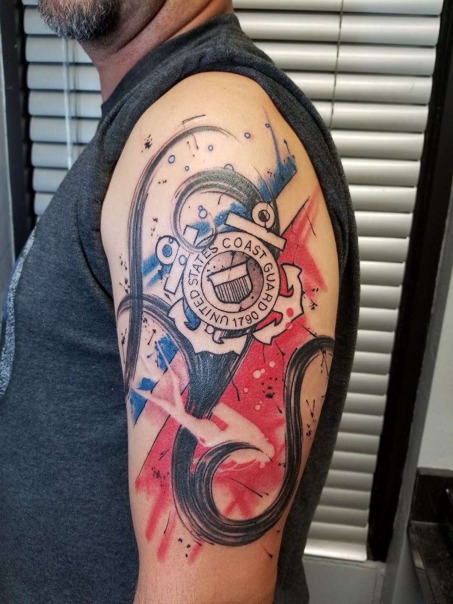 Shoulder tattoo with a circular emblem over red and blue watercolor shapes and black brush strokes