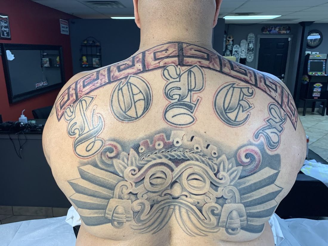 Black and grey upper back tattoo with lettering over an Aztec style sculpted face