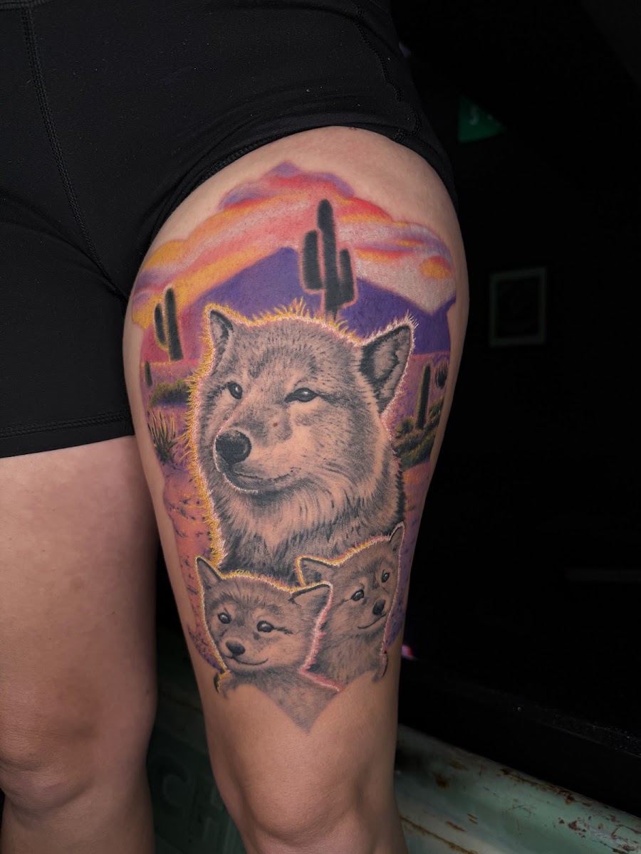 Color realism thigh tattoo of a wolf and two pups in front of a purple Arizona desert sunset with saguaros