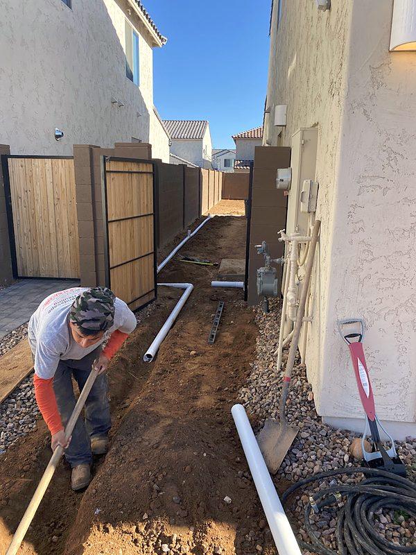A worker digging a trench along a narrow side yard between two stucco walls, white PVC pipe laid beside it