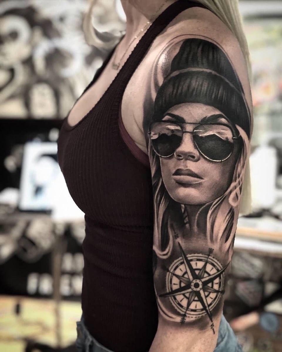 Black and grey portrait tattoo of a woman in sunglasses above a compass on an upper arm