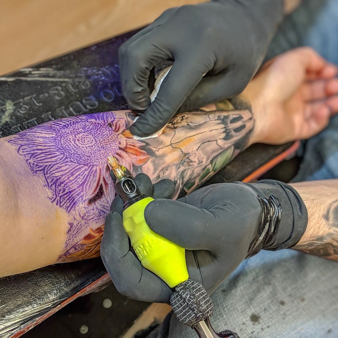 Artist tattooing a forearm over a purple stencil with a yellow grip machine