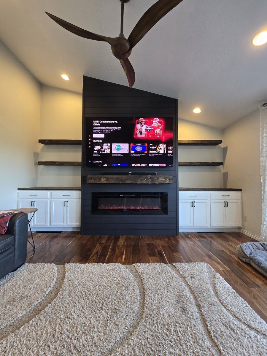 Dark fireplace wall rising to a vaulted ceiling, with TV, electric fireplace, shelves and white cabinets