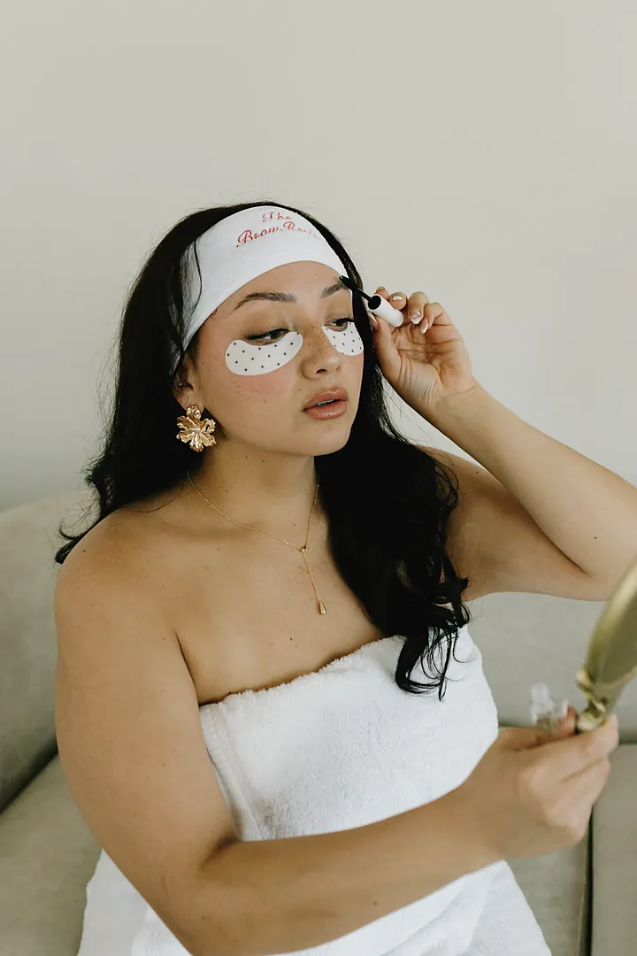 A woman in a white The Brow Recipe headband with dotted under-eye patches brushes her brow, holding a gold hand mirror.