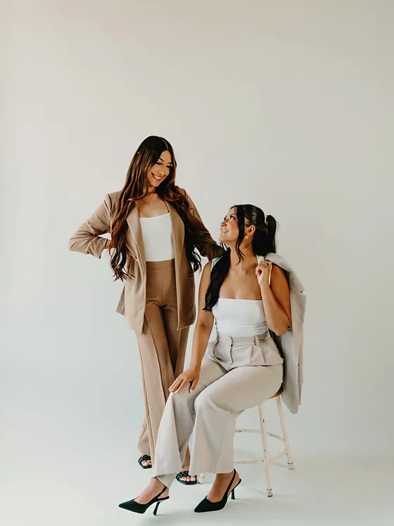 Two Brow Recipe artists in tan and cream outfits posing on a stool against a white wall.