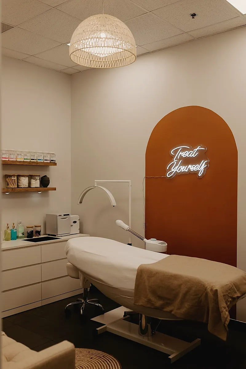 Treatment room with a rust arch wall, a white neon Treat Yourself sign, a rattan pendant lamp and a treatment bed.
