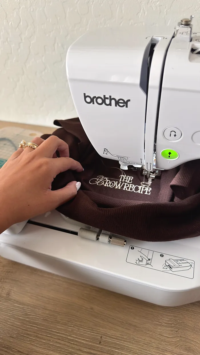 A sewing machine stitching The Brow Recipe wordmark onto a brown shirt.