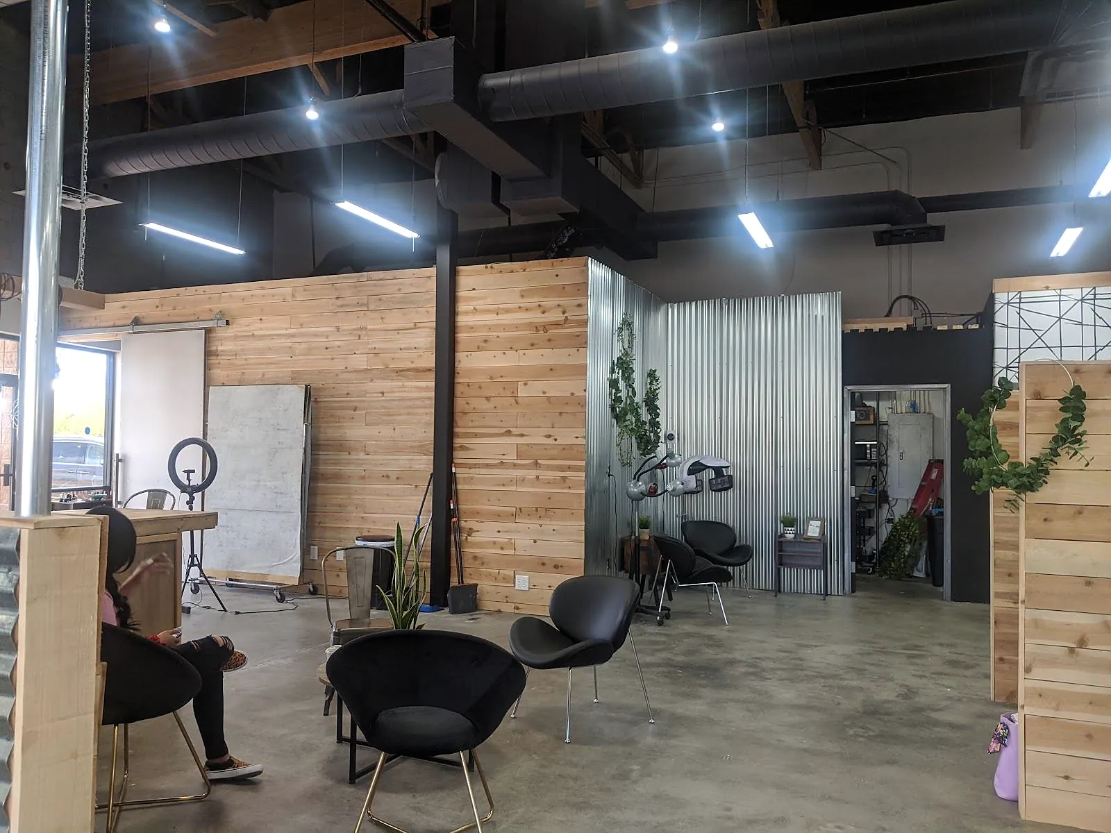 The salon floor: black open ceiling with strip lights, raw pine plank walls, corrugated steel and black chairs on a concrete floor