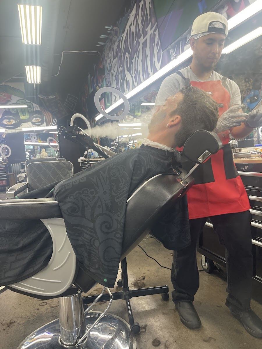 Barber in a red apron giving a client a hot towel steam in a reclined chair at Trnd Setters