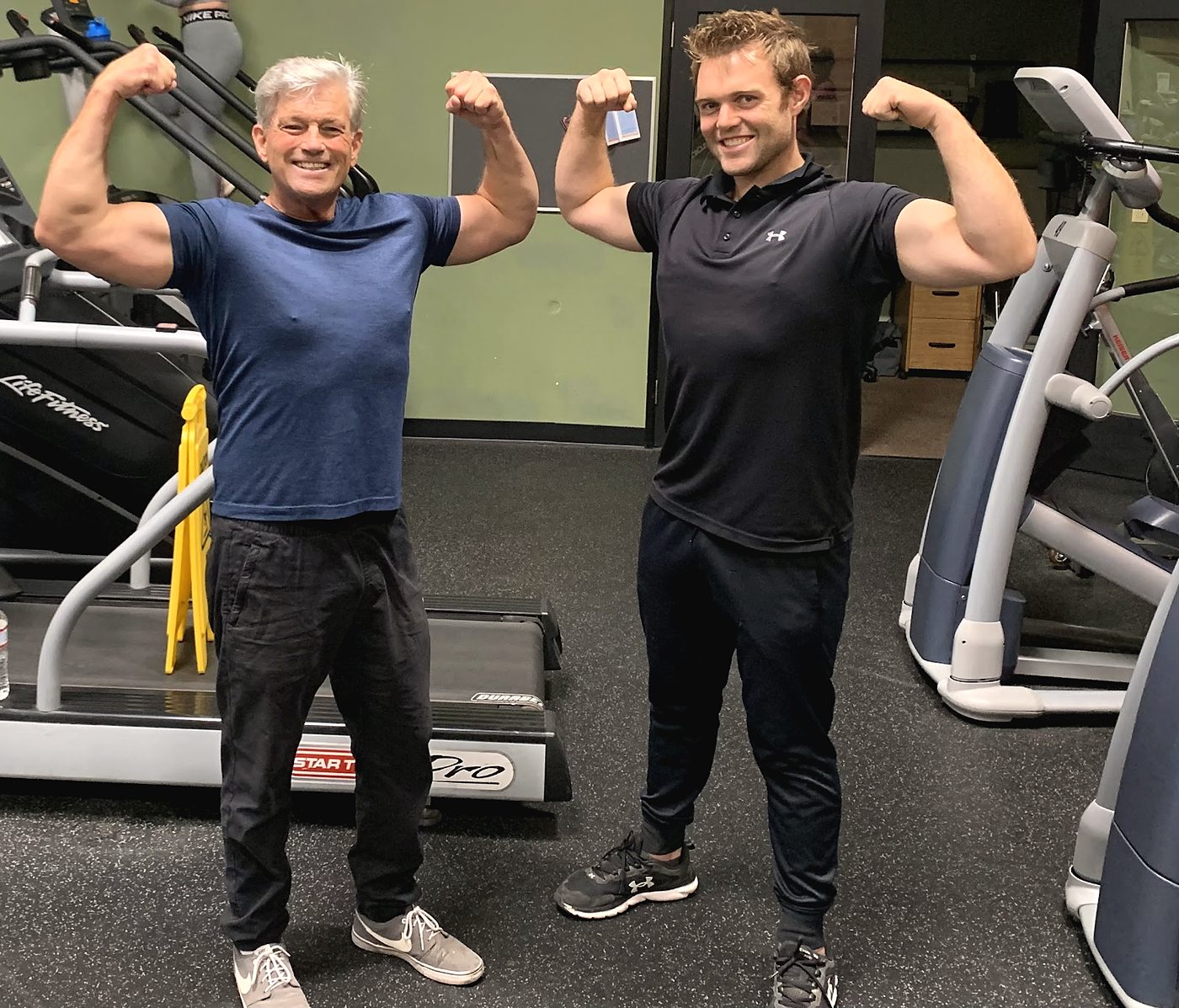 Two men flexing their arms side by side in the gym, a treadmill behind them