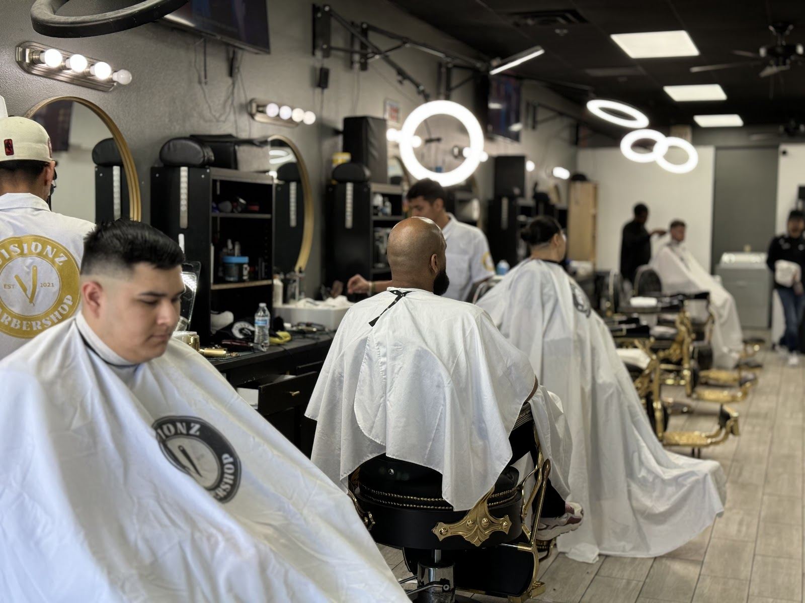 Clients in white Visionz capes at a row of black and gold barber chairs